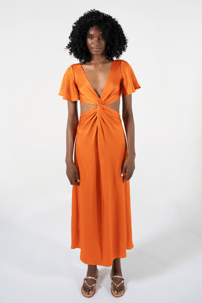 Long orange silk dress with flared short sleeves, low V neck, and a center twist with exposed waist side, worn with gold sandals