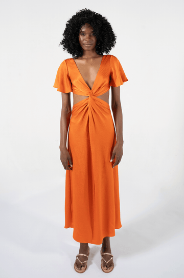 Long orange silk dress with flared short sleeves, low V neck, and a center twist with exposed waist side, worn with gold sandals