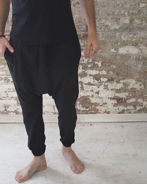 male wearing black harem jogger pants with side pockets, waist elastic, and tapered ankles, worn with black, boat-neck tank top; followed by same white harem jogger pants with relaxed fit, worn with men white scoop neck t-shirt