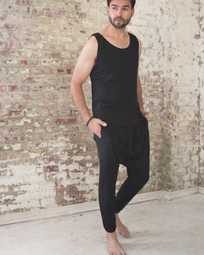 male wearing black sleeveless shirt with wide scoop neck and curved hem. Worn with black harem jogger pants featuring low crotch design, side pockets, waist drawstring and tapered ankles