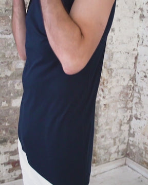 Video of male wearing navy blue hooded aymmetrical shirt, with elbow-length sleeve on one side, sleeveless on the other, and a diagonal hem. Worn with white harem jogger pants featuring waist elastic and drawstring, unique drop crotch, and tapered ankles.